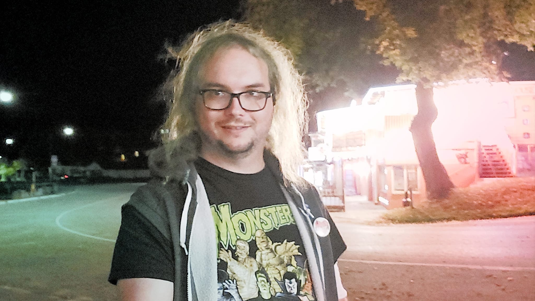 A man with long green hair stands on a street across from a brightly lit bar